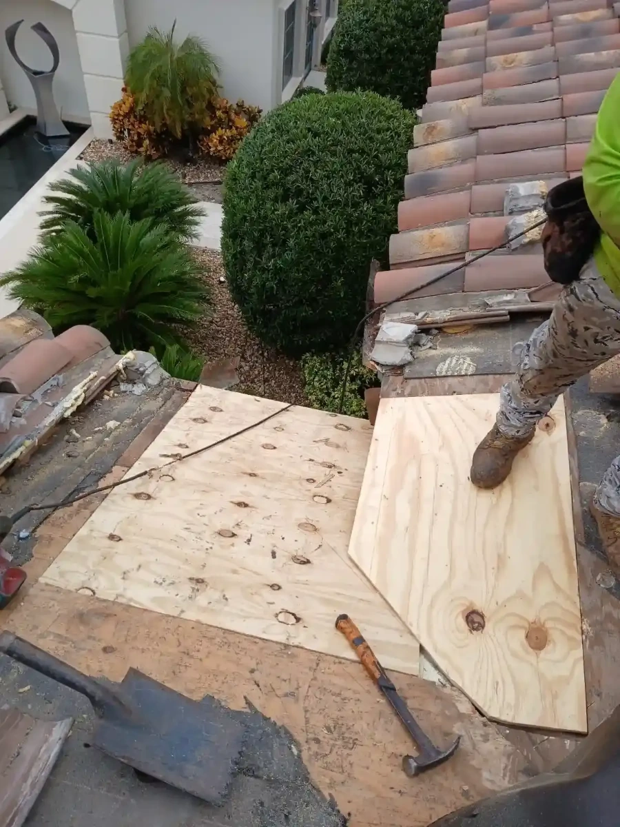 Contractor installing new plywood sheathing sections on Santa Ana tile roof with hammer and fresh lumber replacing rotted decking near chimney area